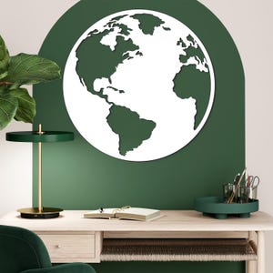 Op de afbeelding: Een wit en groen wandkunstwerk met een wereldkaart. Het kunstwerk is gemonteerd op een groene muur boven een houten bureau. Een groene bureaulamp en een dienblad met kantoorbenodigdheden zijn ook zichtbaar.