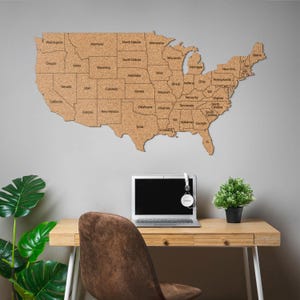 May include: A large, cork map of the United States is mounted on a gray wall. Each state is labeled with its name in a dark font. The map is above a wooden desk with a laptop, headphones, a small plant, and a brown chair.