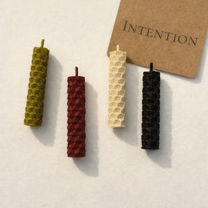 May include: Four pillar candles in olive green, burgundy, cream, and black, each with a honeycomb pattern. A brown tag with the word "INTENTION" is in the background. These decorative candles are ideal for home decor or special occasions.