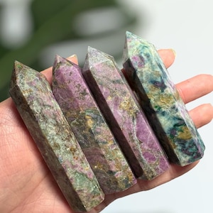 May include: Five green, purple, and gold gemstone points, also known as wands, are arranged in a row on a hand. The points are all approximately the same size and shape.