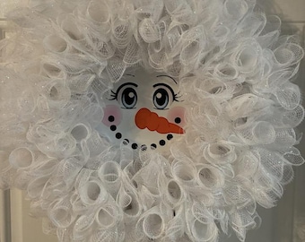 Op de afbeelding: Een grillige sneeuwpopkrans met een lachend gezicht en een wortelneus. De krans is gemaakt van wit gaas en heeft een vrolijk sneeuwpopgezicht met roze wangen en zwarte stipogen. Een feestelijke winterdecoratie.