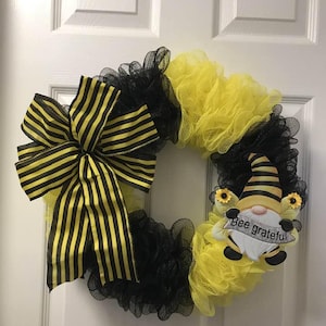 Op de afbeelding: Een zwart-gele bij-thema krans met een zwart-gele gestreepte strik en een bij dwerg met de tekst "Bee grateful".