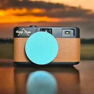 May include: A brown leather camera with a black lens and a turquoise blue lens cap. The camera has the text "Camp Snap" printed on the front.
