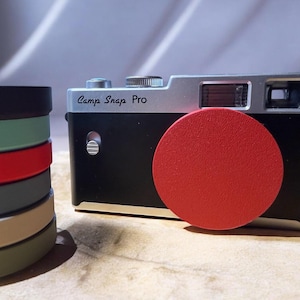 Puede incluir: Una cámara negra y plateada con una tapa de lente roja. La cámara tiene el texto "Camp Snap Pro" impreso en la parte delantera. A la izquierda hay una pila de correas de cámara coloridas en negro, verde, rojo, gris, beige y verde oliva.