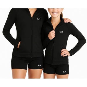 May include: Two people in matching black athletic wear. Each outfit consists of a zip-up jacket and shorts. White initials are embroidered on the jackets and shorts. The outfits are suitable for a team or group.