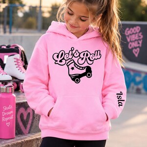 May include: A light pink hooded sweatshirt featuring a black graphic of a roller skate and the text Let's Roll on the chest. The left sleeve is personalised with the name Isla in black script lettering. The hoodie has a front kangaroo pocket.