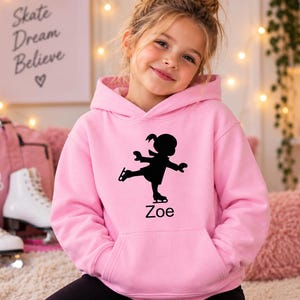 May include: A light pink hooded sweatshirt featuring a black silhouette graphic of a person ice skating. Below the graphic, the name Zoe is printed in black text. The garment includes a front kangaroo pocket and long sleeves with ribbed cuffs.