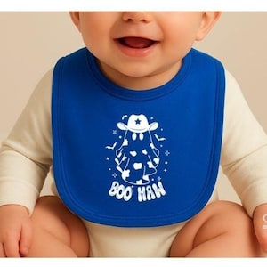 May include: A royal blue baby bib featuring a white graphic print of a ghost wearing a cowboy hat with cow print patterns on its body. The design includes small stars and bats surrounding the ghost, with the text BOO HAW printed below it.