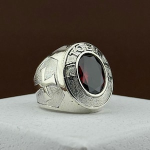 May include: A silver ring with a large, oval, dark red gemstone. The ring has a textured design with the letter "L" on one side and the word "TOB" around the gemstone. The ring is set on a white surface.