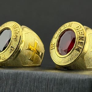 May include: Two gold rings with oval-shaped gemstones. One ring has a dark purple stone, the other a red stone. Both rings feature a cross design and text around the gemstone. The rings are on a dark surface.