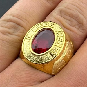 May include: A gold-colored ring with a large, oval, red gemstone at the center. The ring has the words "IN LIEBE DENEN" engraved around the gemstone. The ring is on a finger.