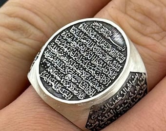 May include: A silver ring with an oval face featuring intricate Arabic calligraphy. The ring has a wide band and a polished finish. The detailed engraving covers the entire face of the ring.