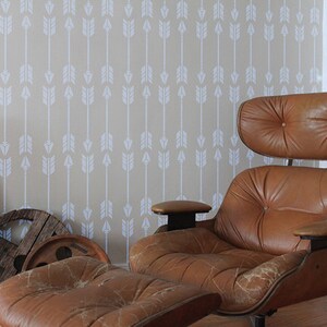 Puede incluir: Un sill&oacute;n y un reposapi&eacute;s de cuero marr&oacute;n de estilo moderno de mediados de siglo. La silla tiene respaldo y asiento acolchados. El fondo presenta una pared beige con un patr&oacute;n de flechas blancas.