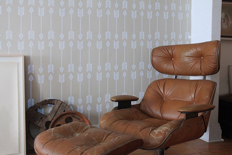 Puede incluir: Un sill&oacute;n y un reposapi&eacute;s de cuero marr&oacute;n de estilo moderno de mediados de siglo. La silla tiene respaldo y asiento acolchados. El fondo presenta una pared beige con un patr&oacute;n de flechas blancas.