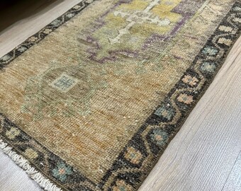 May include: A small, rectangular rug with a faded, vintage look. The rug has a brown, beige, and green color scheme with a geometric pattern in the center. The rug has a fringed edge.