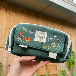 Puede incluir: Un estuche verde con cremallera y ribete blancos. Presenta bordados de flores, mariposas y una ilustración de perro. La palabra "butterfly" está escrita en el estuche.
