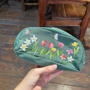 May include: A sage green zippered pouch featuring embroidered floral motifs in shades of white, yellow, pink, and red, accompanied by two small embroidered butterflies in white and purple, set against a textured fabric surface.