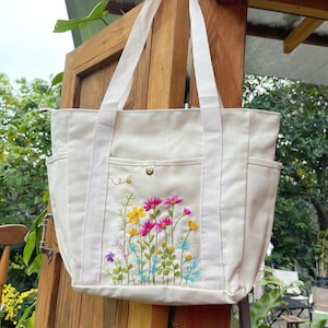 May include: Cream canvas tote bag featuring an embroidered floral design in pink, yellow, blue, and purple. The bag has two external pockets, a snap closure, and white straps. A small embroidered bee is visible.