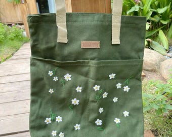 May include: An olive green canvas tote bag with cream-coloured handles and a front pocket. The pocket is decorated with embroidered white daisy flowers with green stems and leaves. A small rectangular patch is sewn on the top.