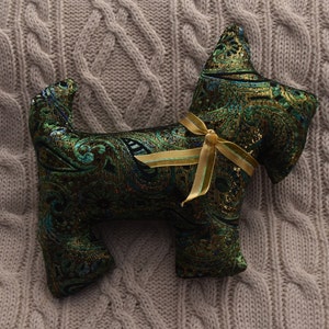 May include: A decorative dog-shaped pillow made of green and gold patterned fabric. The pillow is tied with a gold ribbon. The dog has a paisley-like design and is resting on a gray knitted surface.