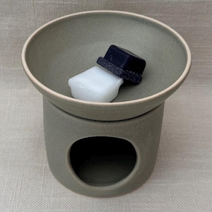 May include: A matte sage green ceramic wax warmer with a wide, shallow bowl top and a circular opening at the base. Inside the bowl sits a two-toned wax melt, featuring a solid white rectangular base topped with a smaller, dark navy blue square.