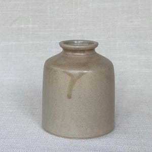 May include: A small, cylindrical ceramic vase with a smooth, matte taupe finish. The vessel features a short, narrow neck with a slightly flared rim and a subtle, darker vertical drip mark running down the upper portion of the body.