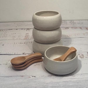May include: Three off-white ceramic bowls stacked on top of each other, with a fourth bowl and a wooden spoon in the foreground. A stack of four small wooden spoons are also visible. The items are on a light-coloured wooden surface.