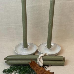 May include: Sage green taper candles with white ceramic candle holders. A bundle of matching candles is tied with a cream ribbon. The arrangement includes a sprig of evergreen and a dried leaf, suggesting a natural theme.