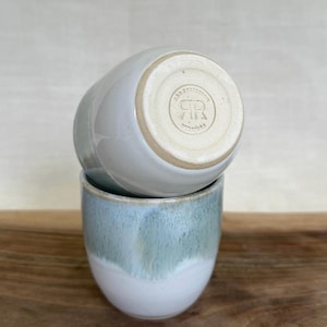 May include: Two ceramic cups, one stacked on top of the other. The bottom cup has a white base with a blue-green glaze that fades towards the top. The top cup is a solid light grey colour. The bottom of the top cup has a stamped logo.