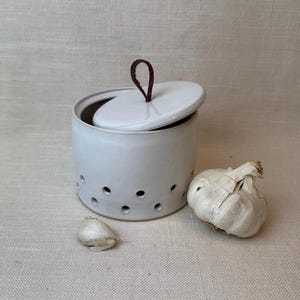 May include: A white ceramic garlic keeper with a lid and a dark brown leather loop handle. The keeper has several small holes around the sides. A whole garlic bulb and a clove are next to the keeper.
