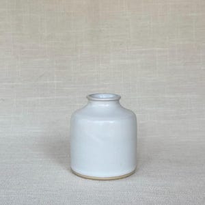 May include: A small, cylindrical ceramic vase with a smooth, matte white glaze. The vessel features a short, narrow neck with a slightly flared rim and a flat base that reveals a thin, unglazed tan ring along the bottom edge of the structure.