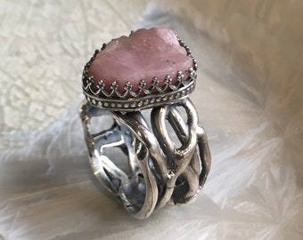 May include: A silver ring featuring a large, rough-cut pink gemstone. The gemstone is set in a crown-like bezel. The ring's band has an intricate, openwork design. The ring is displayed on a white, textured surface.