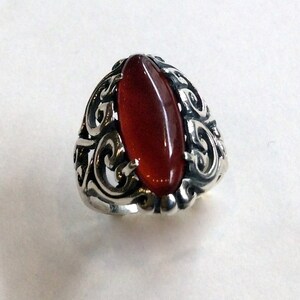 May include: A silver ring with an ornate design featuring a large, oval-shaped carnelian gemstone.