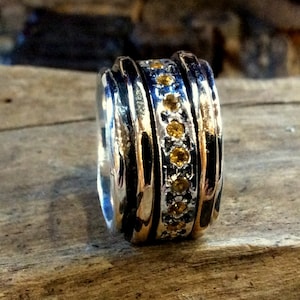 May include: A silver ring with a textured design and a row of small, round, yellow gemstones. The ring has a polished finish and is set against a wooden surface.