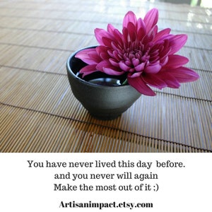 May include: A single pink flower in a black bowl, sitting on a bamboo mat. The text "You have never lived this day before. and you never will again Make the most out of it ;) Artisanimpact.etsy.com" is below the bowl.