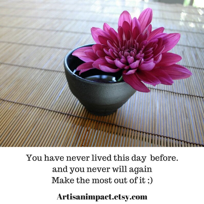 May include: A single pink flower in a black bowl, sitting on a bamboo mat. The text "You have never lived this day before. and you never will again Make the most out of it ;) Artisanimpact.etsy.com" is below the bowl.