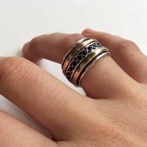 Op de afbeelding: Een zilveren en gouden ring met een band van zwarte edelstenen. De ring wordt aan een vinger gedragen.