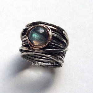 May include: A silver ring featuring a labradorite gemstone set in a rose gold bezel. The ring is crafted from multiple strands of silver wire that are twisted and woven together.