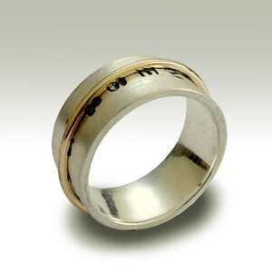 May include: A silver ring with a thin gold band around the edge. The ring has a textured surface and a black inscription.