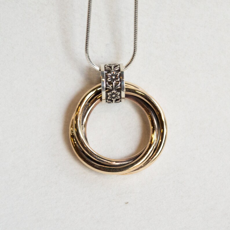 May include: A gold-toned pendant necklace featuring three intertwined rings. The rings are connected to a silver-colored chain through a decorative silver-toned floral bead. The necklace is displayed against a white background.