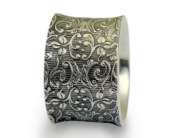 Puede incluir: Un anillo de plata con una banda ancha que presenta un intrincado diseño floral. La banda es texturizada y tiene una forma ligeramente curvada.