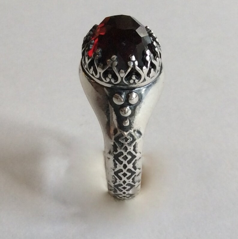 Puede incluir: Un anillo de plata con una piedra preciosa roja engastada en un engaste decorativo en forma de corona. La banda del anillo est&aacute; grabada con un patr&oacute;n geom&eacute;trico.