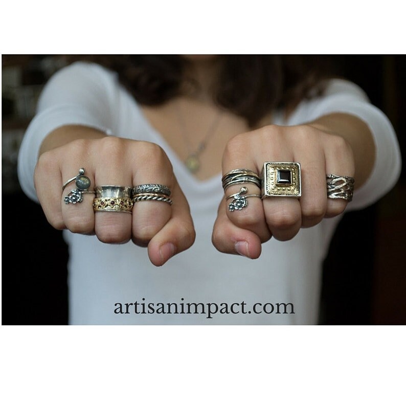 May include: A person's hands wearing a variety of silver and gold rings. The rings feature different designs, including a square ring with a black stone, a ring with a small flower charm, and a ring with a textured band.