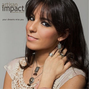 May include: A woman wearing a white lace top with a silver necklace and earrings. The necklace has a square pendant with brown stones. The earrings are silver with a square pendant and brown stones. The text "artisan impact handmade jewelry" and "your dreams miss you" is visible in the image.