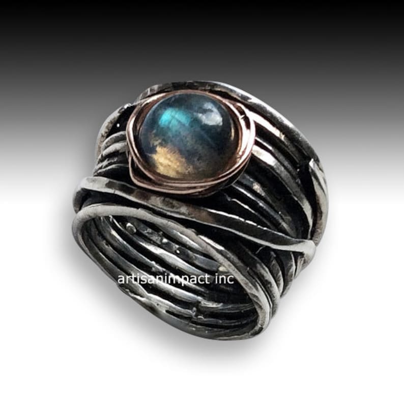 May include: A silver ring featuring a blue and green labradorite gemstone set in a rose gold bezel. The ring is crafted from multiple strands of silver wire that are twisted and woven together.