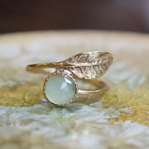 May include: A gold ring with a green gemstone and a leaf design. The ring is on a textured surface.