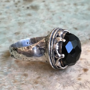Puede incluir: Un anillo de plata con una piedra preciosa negra facetada engastada en un diseño de corona. El anillo está marcado con "925" en la banda interior.