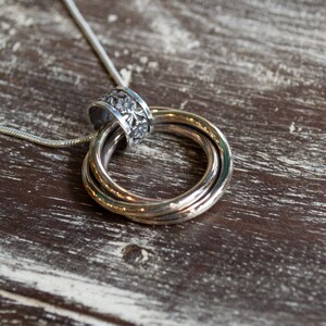 May include: Silver pendant necklace featuring three interlocking rings and a decorative floral-patterned bail. The pendant hangs from a thin, silver chain. The necklace rests on a weathered, wooden surface.
