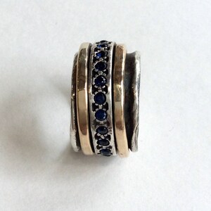 Op de afbeelding: Een zilveren en gouden ring met een band van blauwe saffieren. De ring heeft een getextureerde afwerking en is ontworpen om te draaien.