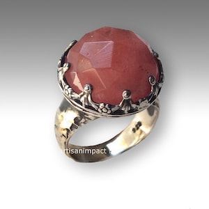 Puede incluir: Un anillo de plata con una gran piedra preciosa rosa facetada engastada en un engaste detallado y ornamentado en forma de corona.